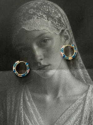 Blåa creoler med blommönster - Coola örhängen i creolmodell med blå bas och färgglada blommor i grönt, rosa och vitt. Örhängena har en rund form och detaljer i guldfärg. Perfekta för dig som vill sticka ut med färg och mönster. Materialet ser ut att vara metall med emalj.