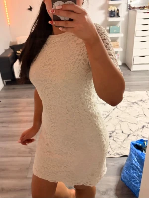 Vit spetsklänning med kort ärm - Säljer en vit, figurnära spetsklänning med korta ärmar och rund hals. Klänningen har ett vackert blommigt spetsmönster och är i kort modell. Perfekt för sommarens alla tillfällen!