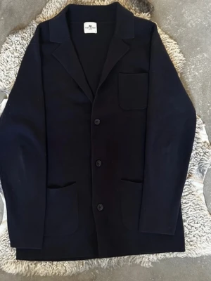 Mörkblå kavaj från John Henric - Stilren mörkblå kavaj från John Henric med tre knappar framtill och tre utanpåliggande fickor. Klassisk krage och rak passform. Perfekt för en clean och modern look. Materialet ser ut att vara mjukt och bekvämt, troligen bomull eller ullblandning.