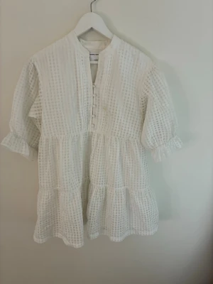 Vit kortklänning med volang - Söt vit kortklänning i luftig bomull med rutigt strukturmönster, volangdetaljer och knappar framtill. Klänningen har trekvartsärm med rynk vid ärmslut och avslappnad passform. Perfekt för varma dagar och enkel att styla.