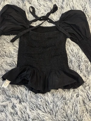 Svart topp med puffärmar och volang - Trendig svart topp med smockad ( snatcing) kropp, stora puffärmar och volangkant nedtill. Toppen har knytband vid axlarna där bak och är tillverkad i ett lätt tyg som ger en snygg siluett. Märket är missguided, det står storlek 40 men skulle säga att den är väldigt liten i storlek och är mer storlek S. 