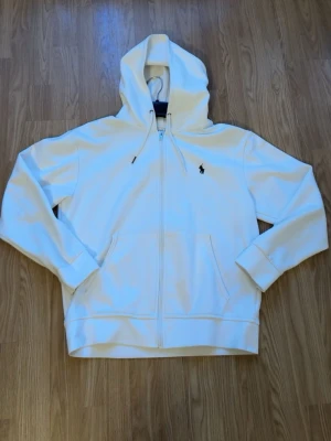 Vit hoodie från Polo Ralph Lauren - Snygg vit hoodie med dragkedja från Polo Ralph Lauren. Klassisk design med huva, känguruficka och den ikoniska broderade loggan på bröstet. Perfekt för en clean och stilren look. Dock så saknar den metallbiten på ena snöret. Hör av dig via intresse