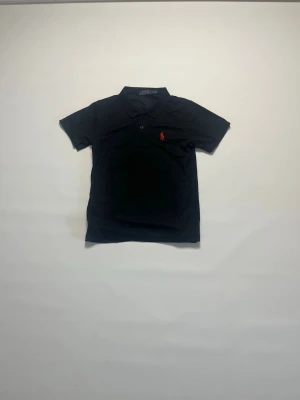 Svart pikétröja från Ralph Lauren storlek S - Klassisk svart pikétröja från Ralph Lauren med korta ärmar och krage. Tröjan har en röd broderad logga på bröstet och knäppning framtill. Perfekt för en stilren och sportig look. Den är i storlek S och är helt ny och aldrig buren