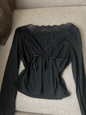 Svart långärmad topp med spets - svart topp med långa ärmar och djup v-ringning dekorerad med spets. Blusen har en feminin passform med knytband i midjan och är gjord i ett mjukt, stretchigt material. Perfekt för dig som gillar en stilren men ändå romantisk look.💞💞💕