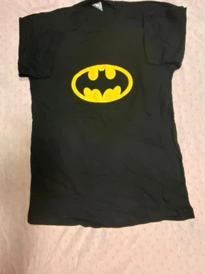 Svart Batman t-shirt  - Cool svart t-shirt med det klassiska Batman-logotypen i gult på bröstet. Perfekt för dig som gillar superhjältar och vill ha en enkel men snygg look. T-shirten har korta ärmar och rund hals. Materialet känns mjukt och bekvämt.  Helt ny 
