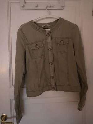 Beige jeansjacka från Taifun - Säljer en grön jeansjacka från Taifun med rund krage och dekorativa sömmar. Jackan har knappar framtill och två bröstfickor med lock och knapp. Perfekt för dig som vill ha en stilren och enkel look.