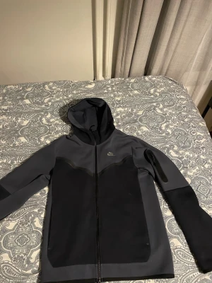 Svart och mörkgrå hoodie från Nike - Cool hoodie från Nike i svart och mörkgrått med hel dragkedja och huva. Materialet är slätt och stretchigt, perfekt för en sportig look. Nike-loggan syns diskret på bröstet. Långärmad och med snygga paneler på framsidan. Använt en gång