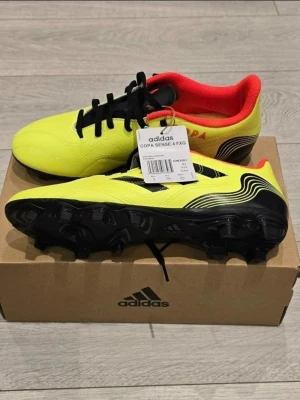 Adidas Copa Sense.4 FXG fotbollsskor - Snygga fotbollsskor från Adidas i modellen Copa Sense.4 FXG. Skorna är gula med svarta detaljer och har en röd insida. De har klassisk snörning och yttersula med fasta dobbar för spel på fasta underlag. Tillverkade i syntetmaterial för bra komfort och hållbarhet.