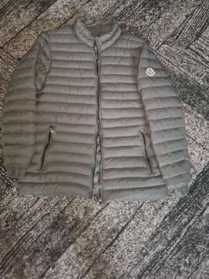 Grå dunjacka från Moncler - Snygg grå dunjacka från med quiltad design och två dragkedjeförsedda fickor framtill. Jackan har hög krage och på ärmen. Perfekt för kalla vinterdagar och riktigt stilren look.