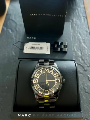 Svart och guld klocka Marc Jacobs - Stilren klocka från Marc by Marc Jacobs i svart metall med guldfärgade detaljer. Klockan har en rund urtavla med stora bokstäver runt kanten och länkarmband. Perfekt accessoar för att lyfta din outfit.