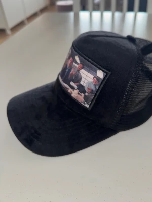 Giovinz keps med wolf of wallstreet motiv - Svart truckerkeps med böjd skärm och mesh på sidorna. Framtill finns ett tygmärke med ett färgglatt motiv av flera personer. Kepsen har justerbar snapback baktill och är tillverkad i polyester och mesh. Perfekt för en avslappnad streetstil.