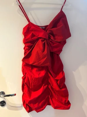 Röd kort klänning med rosett - Röd kort klänning med smala axelband och stor rosett framtill. Draperad och voluminös design som ger en cool och unik look. Klänningen är ärmlös och har en tight passform upptill med volangdetaljer nedtill. Perfekt för dig som vill sticka ut.