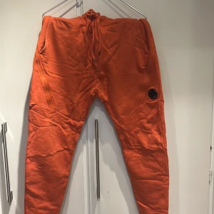 Orange mjukisbyxor från C.P. Company - Säljer ett par orange mjukisbyxor från C.P. Company med dragsko i midjan och ribbade muddar vid bensluten. Byxorna har en unik detalj med en svart lins på ena benet och är tillverkade i mjukt bomullsmaterial. Perfekta för en chill och avslappnad stil.