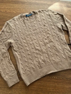 Beige kabelstickad tröja Polo Ralph Lauren - Kabelstickad beige tröja från Polo Ralph Lauren med rund halsringning och ribbade muddar. Klassisk design med broderad logga på bröstet. Perfekt för lager-på-lager och chill stil. Nästan oanvänt och inga fläckar. Storlek M, pris kan diskuteras