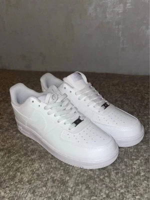 Nike Air Force 1 vita sneakers - Säljer ett par klassiska Nike Air Force 1 sneakers i helvitt skinn med snörning. Modellen har en chunky sula och perforeringar på tån för extra ventilation. Perfekt för dig som vill ha en clean och tidlös look. storlek 42-43-44