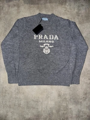 Grå stickad tröja från Prada - Snygg grå stickad tröja från Prada med vit logga och texten 'PRADA MILANO' på bröstet. Tröjan har rund halsringning, ribbade muddar och nederkant. Perfekt för dig som gillar exklusiva och trendiga plagg.