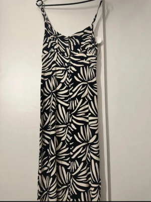 Svartvit långklänning med mönster - Cool långklänning med svartvitt bladmönster och smala axelband. Klänningen har en snygg passform och knappar framtill. Perfekt för dig som gillar att sticka ut med ett grafiskt mönster. Materialet känns lätt och flowy, perfekt för varma dagar.