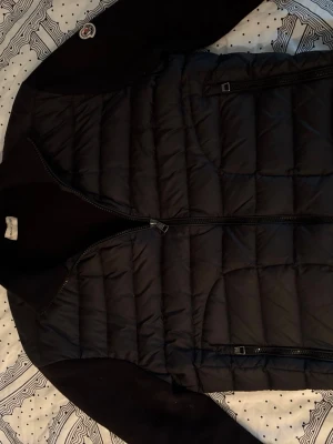 Svart cardigan från Moncler - Snygg svart cardigan från Moncler med quiltat mönster och dragkedja framtill. Västen har Moncler-logga på axeln och är tillverkad i ett glansigt material med klassisk passform. Perfekt för lager-på-lager och streetstyle. Även perfekt nu när solen tittar fram!