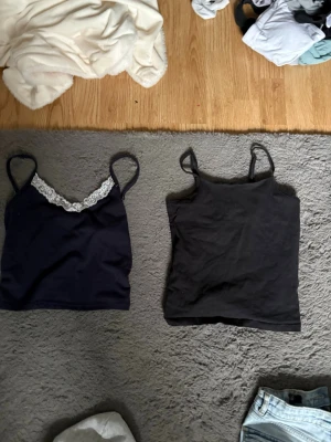 Två svarta linnetoppar - Säljer två svarta linnetoppar med smala axelband. Ena toppen har en snygg spetsdetalj vid urringningen och den andra är enkel och stilren. Perfekta att matcha med jeans eller kjol för en trendig look. Det med spets är i xxs och det andra är i 140🫶🏻💕