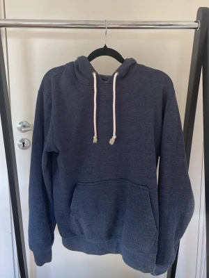 Mörkblå hoodie med vita snören - En stilren mörkblå hoodie med klassisk känguruficka och vita dragsnören i huvan. Hoodien har en avslappnad passform och är tillverkad i mjukt bomullsmaterial. Perfekt för en chill och casual look.