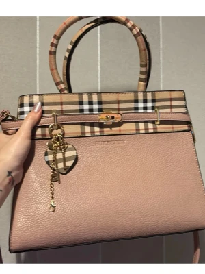 Burberry rosa handväska med rutigt mönster - Säljer en snygg rosa handväska från Burberry med klassiskt beige, svart och rött rutmönster upptill och på handtagen. Väskan har guldfärgade detaljer och en matchande nyckelring med hjärta. Tillverkad i strukturerat läder.