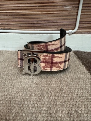 Rutig Burberry bälte med B-spänne - Snyggt bälte från Burberry med klassiskt rutigt mönster i beige och bruna toner. Bältet har ett stort metallspänne med ett B framtill och detaljer i skinn längs kanterna. Perfekt accessoar för att lyfta din outfit.