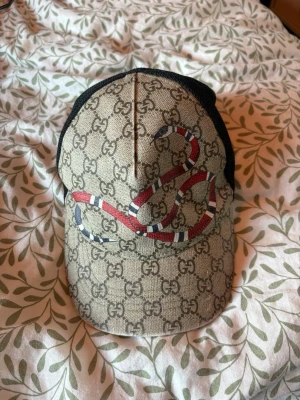 Gucci keps med orm och GG-mönster - Snygg keps från Gucci med klassiskt GG-mönster i beige och brunt, prydd med en röd och vit orm på framsidan. Baksidan är i svart mesh med justerbart band i grönt och rött. Perfekt accessoar för en trendig streetstyle-look.