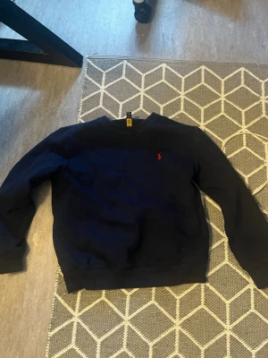 Ralph Lauren sweater - Säljer en mörkblå sweatshirt från Polo Ralph Lauren med klassisk rund hals och röd broderad logga på bröstet. Tröjan har långa ärmar och ribbade muddar vid ärmslut och nederkant. Perfekt för en stilren och avslappnad look.