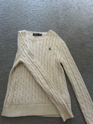 Beige stickad tröja från Polo Ralph Lauren - Kabelstickad beige tröja från Polo Ralph Lauren med rund halsringning och lång ärm. Tröjan har det klassiska broderade logotypen i mörkblått på bröstet och ribbade muddar vid ärmslut och nederkant. Perfekt för lager-på-lager och snygg till jeans. Storlek S och sitter super fint, nästan aldrig använd. 