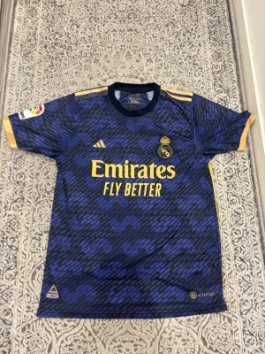 Real Madrid bortatröja Vini Jr #20 - Snygg Real Madrid tröja i mörkblått med gula detaljer och tryck. Tröjan har Vini Jr och nummer 20 på ryggen, samt klubbmärke och sponsorlogga på bröstet. Tillverkad i lätt och ventilerande material, perfekt för fotbollsträning eller match.