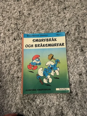 Smurfbråk och Bråksmurfar bok - Smurfarnas äventyr: Smurfbråk och Bråksmurfar är en färgglad seriebok med blå, röda och vita illustrationer på omslaget. Boken är i mjukt papper och har tecknade smurfar på både fram- och baksida. Perfekt för dig som gillar klassiska serier och humor.