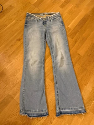 Ljusblå bootcut jeans med råa kanter - Säljer ett par ljusblå bootcut jeans med slitningar och råa, fransiga kanter både i midjan och vid bensluten. Jeansen har klassiska fem fickor och en avslappnad vintage vibe. Perfekta för dig som gillar en retro och avslappnad stil.