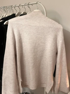 Ljus stickad polotröja - Mysig ljus stickad polotröja med ribbad struktur och långa ärmar. Tröjan har en hög krage och slitsar i sidorna för en avslappnad look. Perfekt att styla med jeans eller kjol.