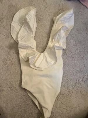 Vit body med volangdetaljer - Säljer en snygg vit body med djup urringning och stora volangdetaljer vid axlarna. Bodyn har smala axelband och är tillverkad i ett mjukt, stretchigt tyg som sitter skönt mot kroppen. Perfekt för dig som vill ha något extra till outfiten. Från Zara i storlek S men men skulle säga att den sitter som en xs!!💕