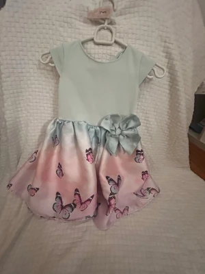 Pastellfärgad klänning med fjärilar - Supersöt klänning i pastellfärger med ljusblå överdel och kjol i rosa och blå toner dekorerad med fjärilar. Kjolen har en stor rosett i midjan och klänningen har korta ärmar. Perfekt för dig som gillar gulliga detaljer och mjuka färger. Storlek vet ej passar mellan 5-7 år ungefär 