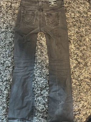 Grå bootcut jeans från True Religion - Säljer ett par gråa bootcut jeans från True Religion med klassiska kontrastsömmar och broderade bakfickor. Jeansen har fem fickor, normal midja och är tillverkade i ett mjukt denimtyg. Defekterna är att dragkedjan är sliten och är lite krånglig men den funkar som man ser på bilden, samt slitet på nedre delen av högra benet.