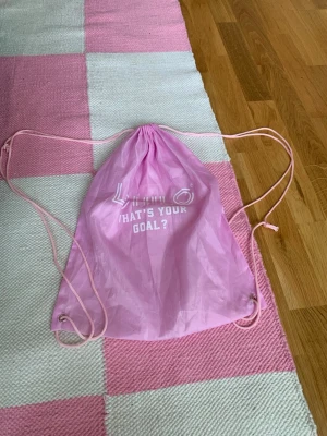 Rosa gympåse från Lohilo - Säljer en ljusrosa gympåse från Lohilo med vitt tryck där det står 'WHAT'S YOUR GOAL?'. Påsen har tunna snören i samma färg och metallöljetter i hörnen. Perfekt för träning eller att bära med sig det viktigaste. Har lagat en söm för hand och metallringar är slitna.
