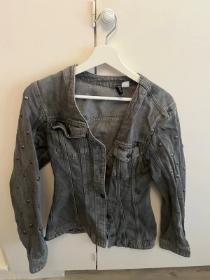 Grå jeansjacka med nitar från H&M - Snygg grå jeansjacka från H&M med coola nitar längs ärmarna. Jackan har klassisk knappstängning framtill, två bröstfickor med lock och markerade sömmar. Perfekt för dig som vill ha en edgy och trendig look.
