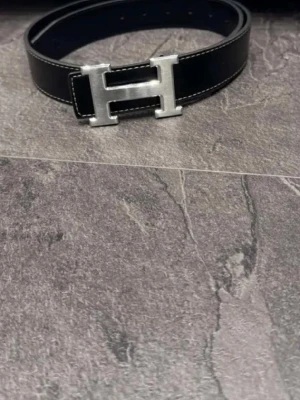 Svart  skärp med H-spänne - Snyggt svart skärp från  med det ikoniska H-spännet i silverfärgad metall. Skärpet är gjort i skinn och har vita sömmar längs kanterna för en stilren kontrast. Perfekt accessoar för att lyfta vilken outfit som helst.