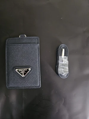 Svart korthållare - Stilren svart korhållare i strukturerat läder med ikonisk metallogo framtill. Kommer med tillhörande svart rem med silverfärgad detalj. Perfekt för kort och ID, smidig att bära runt halsen eller i väskan.