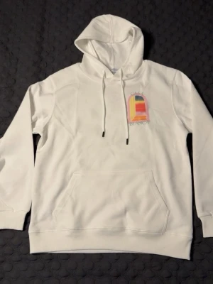 Vit hoodie från Casablanca - Snygg vit hoodie från Casablanca med färgglad grafisk print och texten 'Casa Blanca' på både bröstet och ryggen. Hoodien har huva med dragsko och en stor magficka. Perfekt för dig som gillar streetwear och stilrena detaljer. STORLEK M, PASSAR S OCH M. HELT NY, OANVÄND!