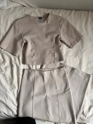 Beige set med topp och tjol - Snyggt tvådelat set från Gina tricklt  i beige nyans. Setet består av en kortärmad topp med rund hals och raka linjer samt matchande höga tjol med pressveck. Materialet är mjukt och stretchigt för en bekväm passform. Perfekt för en stilren och modern look.