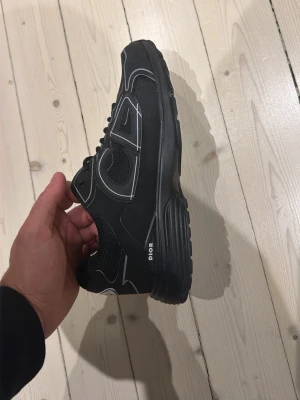 Dior B30 Triple Black - Snygga svarta sneakers  med vita kontrastdetaljer och logga på sidan. Skorna har en sportig siluett, svarta skosnören och en robust sula. Materialet är en mix av mesh och syntet för en modern look.