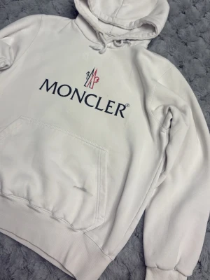 Vit Moncler hoodie med logga - Snygg vit hoodie från Moncler med stor svart logga och röd detalj framtill. Klassisk känguruficka och huva med dragsko. Mjuk bomullskänsla och ribbade muddar vid ärmslut och nederkant. Perfekt för en clean och stilren look. Finns lite skavanker på olika ställen. Kolla noga på bilderna där allt dokumenterats. 