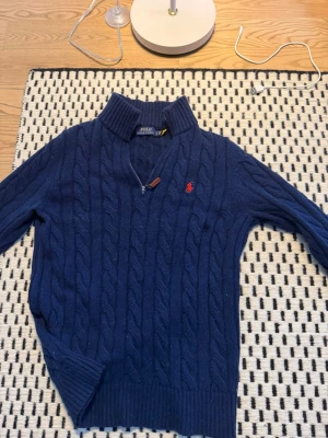 Blå kabelstickad tröja Polo Ralph Lauren - Mörkblå kabelstickad tröja från Polo Ralph Lauren med halv dragkedja och ribbad hög krage. Tröjan har det klassiska röda Polo-logot broderat på bröstet och ribbade muddar vid ärmslut och nederkant. Perfekt för lager-på-lager-stil.