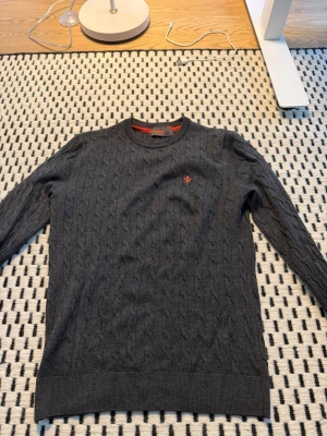 Mörkgrå kabelstickad tröja Gant - Mörkgrå kabelstickad tröja från Gant med rund halsringning och diskret röd logga broderad på bröstet. Tröjan har långa ärmar och ribbade muddar vid ärmslut och nederkant. Perfekt för lager-på-lager och klassisk stil. Tröjan har tre pytte pytte pytte små hål kan skicka bild ifall ni är intresserade.