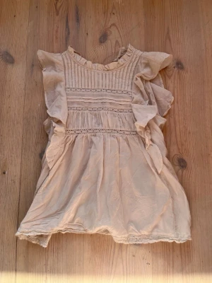 Beige volangklänning med spetsdetaljer - Supersöt beige klänning med volanger på axlarna och fina spetsdetaljer framtill. Klänningen har rund halsringning med liten volangkant och är ärmlös. Perfekt för sommarens alla tillfällen!