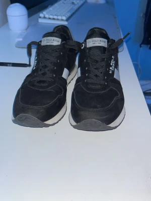 Svarta skor från U.S. Polo Assn. - Säljer ett par snygga svarta sneakers från U.S. Polo Assn. med vita detaljer och logotyp på sidan. Skorna har klassisk snörning, vit sula och är tillverkade i en kombination av mocka och textil. Perfekta för dig som vill ha en stilren och sportig look.