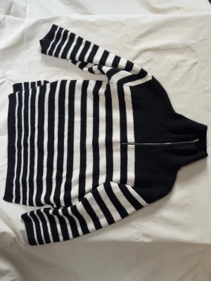 Randig stickad tröja från H&M - Säljer en svartvit stickad tröja från H&M med breda och smala horisontella ränder. Tröjan har halv dragkedja vid halsen och ribbstickad struktur. Hög krage och passform.