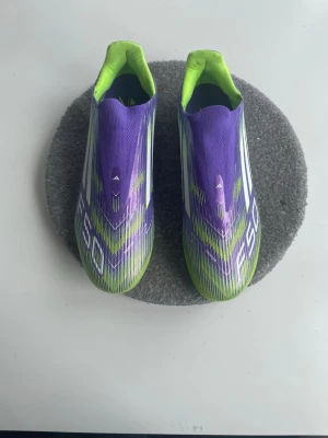 Adidas F50 lila och lime fotbollsskor - Säljer ett par Adidas F50 fotbollsskor i en snygg lila och limegrön färgkombination. Skorna har en strömlinjeformad design med vita detaljer och klassiska Adidas-ränder på sidan. Ovandelen är i syntetmaterial för lätt vikt och bra bollkontroll. Och har använts bara en gång 